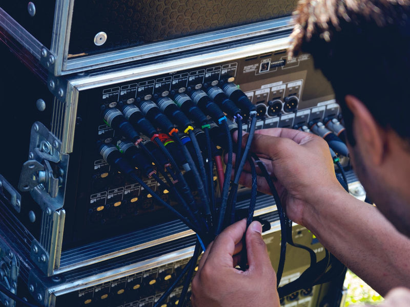 A close-up of a person's hands connecting multiple black audio or cable ties to a rack of electronic equipment with metal protective cases.